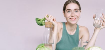 Woman in tank top smiling with broccoli, cabbage, and glass of water on table.