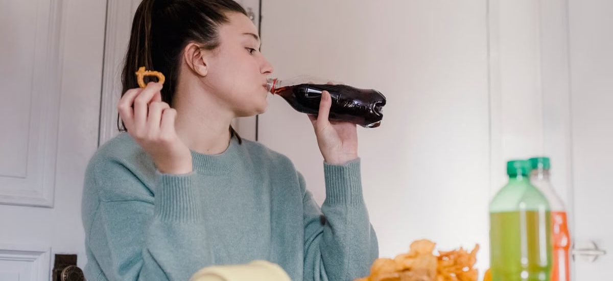 Young female drinking sweet tasty lemonade from plastic bottle at table with different snacks