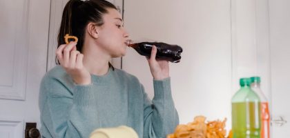 Young female drinking sweet tasty lemonade from plastic bottle at table with different snacks