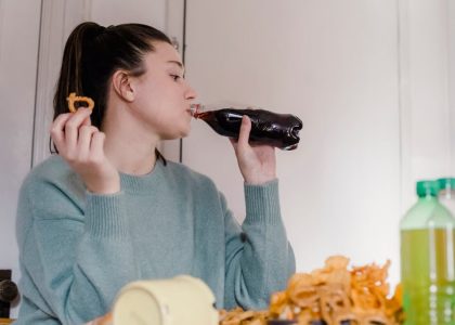 Young female drinking sweet tasty lemonade from plastic bottle at table with different snacks